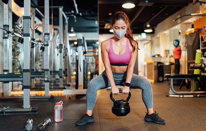 a masked woman who has covid doing kettlebell squats in a gym
