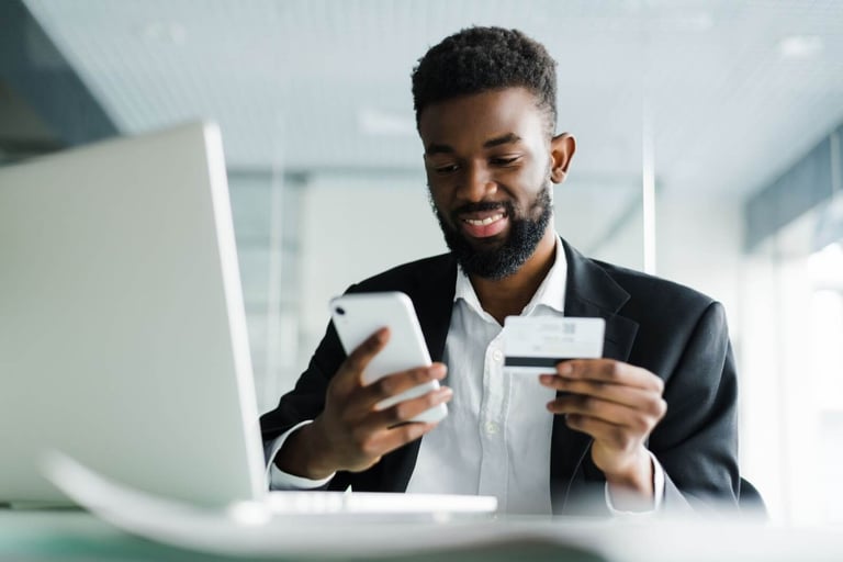 Imagem de um homem negro segurando um cartão e um celular. Ele está sentado à frente de um notebook branco. O homem está de terno e sorri para o celular. O fundo é branco e está embaçado