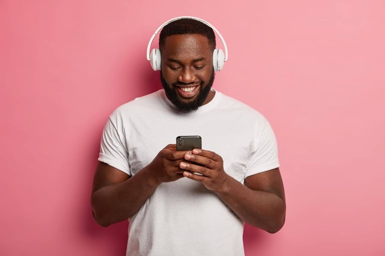Um homem negro, de barba, com camiseta branca e fones de ouvido branco, está sorrindo enquanto olha para o celular