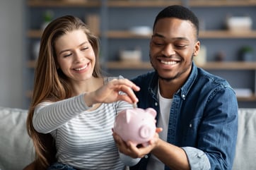 Family Savings. Young Interracial Couple Putting Coin In Piggybank At Home