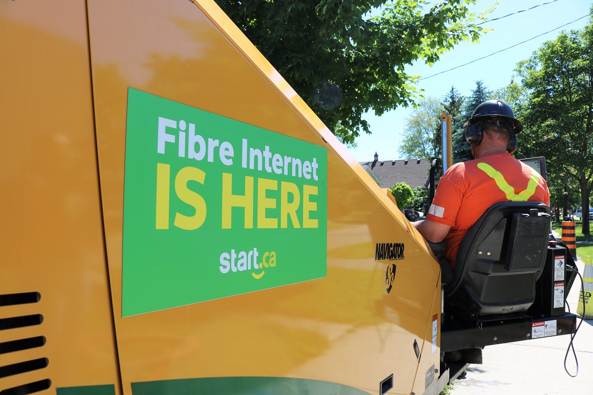 In  the foreground pictures a closeup of a yellow construction machine. On the side surface of the machine is a large green sign that says "Fibre internet is here - Start.ca". On the right hand side of the image pictures a construction worker, operating the machine. 