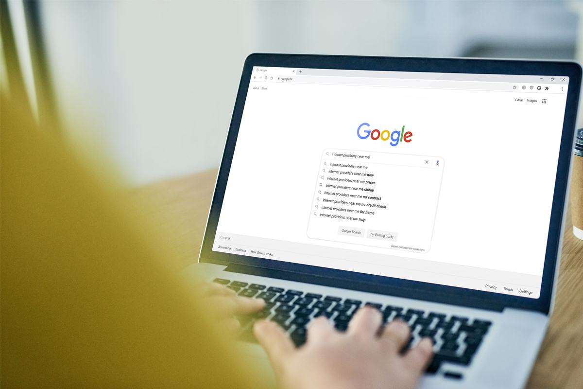 A laptop screen open to the google webpage in the middle of a search with a drop down list of results. In the foreground is a blurred arm on the left hand-side of the picture. Hands type on the laptop keyboard. 