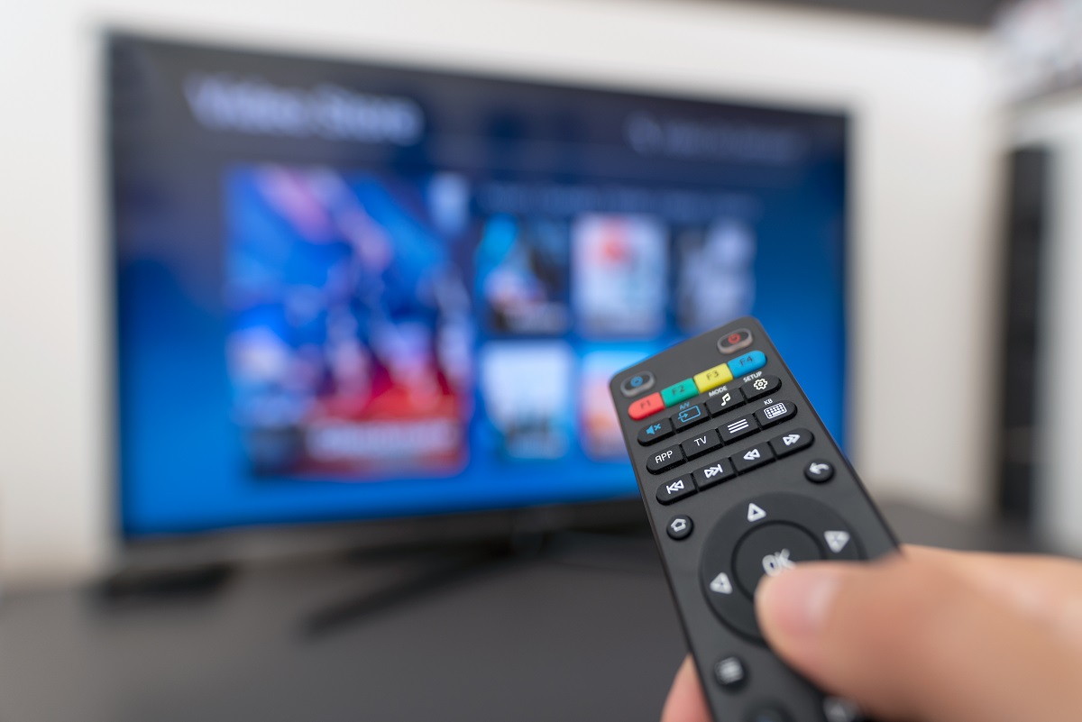 Foreground features partial hand holding a black remote. The thumb is over the down arrow on the remote. In the background is a powered on TV that is blurred. 