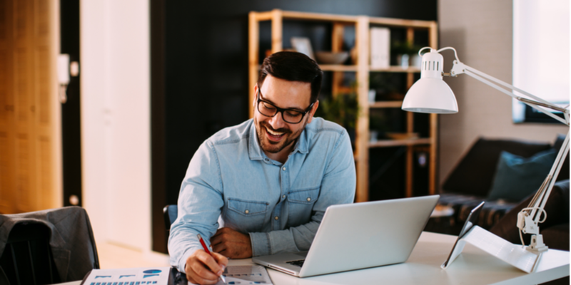 business plan for working remotely Man in glasses sitting at desk in front of a laptop while writing into an open notebook