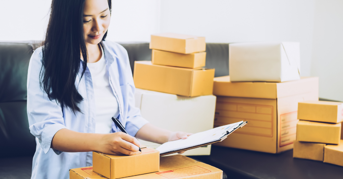 Woman surrounded by boxes holding a check list and writing something down with a black pen