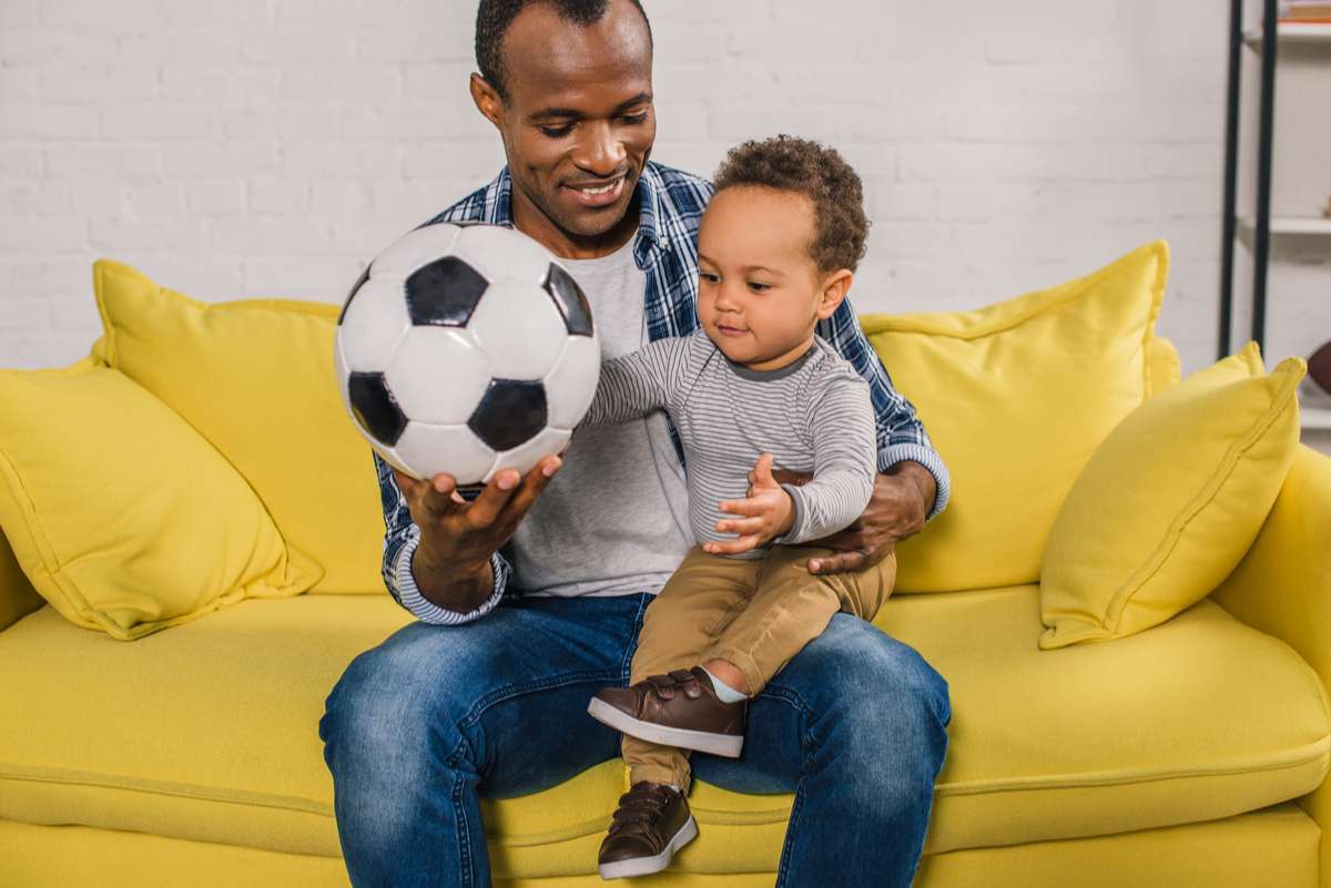 Happy young father holding soccer ball while sitting with adorable little son on sofa (R) (S)