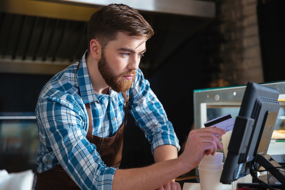 Handsome concentrated bearded waiter swiping credit card through the computer terminal in cafe.jpeg