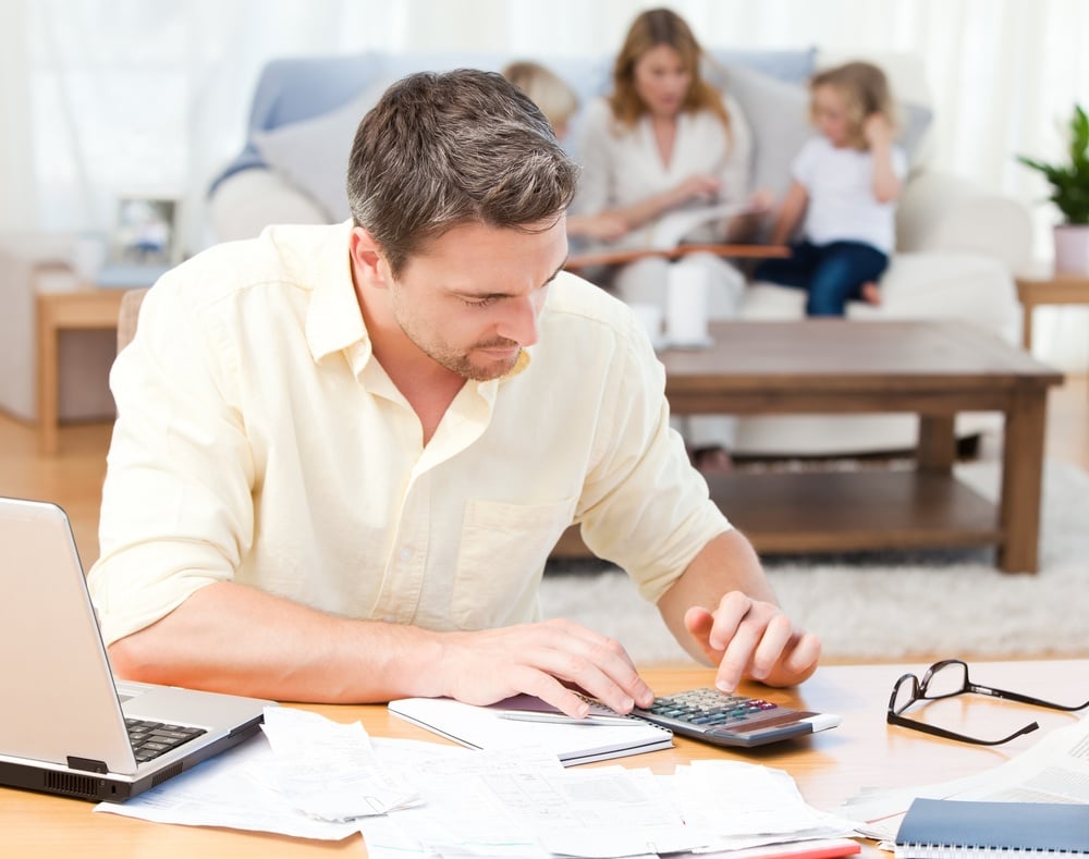 Man calculating his bills while his family are sitting on the sofa