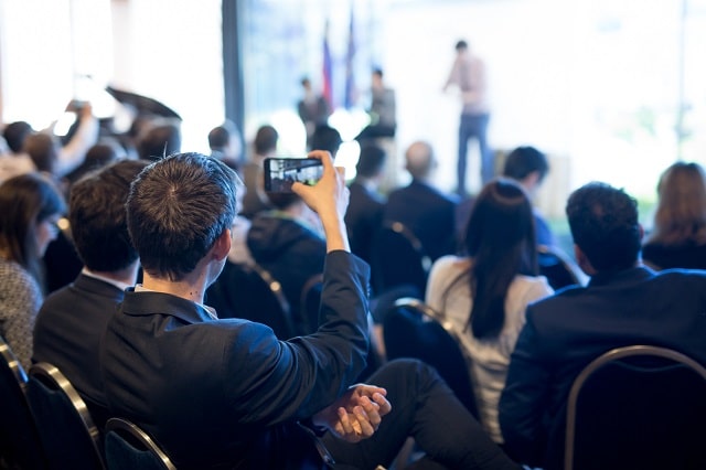 27961394_businessman-takes-a-picture-of-corporate-business-presentation-at-conference-hall-using-smartphone-min